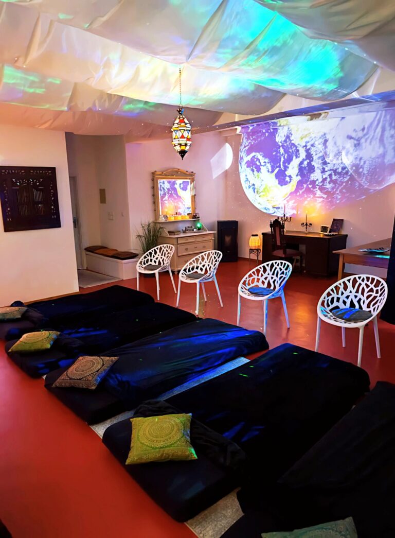 Das Bild zeigt den Übungsraum für die Atemmeditation Lünen. Es stehen vier Stühle bereit und vier Matten zur Entspannung. Der Raum hat einen roten Fußboden. Die Wände werden mit Lichtmotiven angestrahlt, die Decke ist mit Tüchern abgehängt.